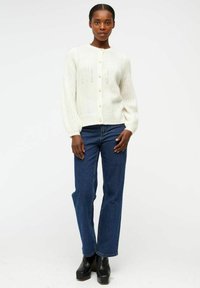 Cardigan en tricot blanc avec un col rond, fermeture à boutons et motif texturé. Associé à un jean évasé taille haute bleu et des bottes à plateforme noires.