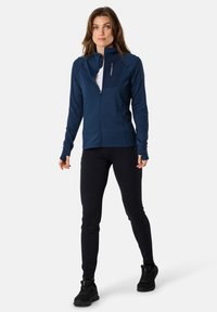 Marinblå zip-up jacka med hög krage och texturerade paneler, ihop med svarta leggings och svarta träningsskor. Slät tyg, elegant design.
