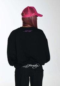 Felpa nera oversize con dettaglio logo viola sul retro, abbinata a pantaloni neri con logo ricamato bianco sulla vita. Cappellino rosa.