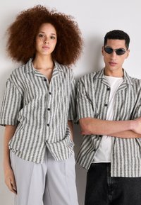Le camicie a maniche corte con bottoni presentano un motivo a righe in grigio e bianco. Il modello maschile indossa occhiali da sole; entrambi hanno una vestibilità rilassata.