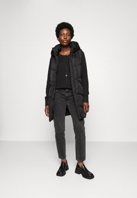 Gilet matelassé noir avec capuche, porté sur un pull en tricot noir et un jean gris. Des chaussures à talons carrés noires complètent la tenue.