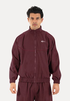 Hombre vestido con una chaqueta granate con cremallera y pantalones a juego con puños elásticos, de pie frente a un fondo claro y liso.