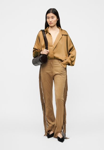 Καφέ σατέν μπλούζα με βαθύ V-λαιμόκοψη, συνδυασμένη με καφέ παντελόνια φαρδιάς γραμμής με λεπτομέρειες από φούντες. Το μοντέλο κρατά στρογγυλή μαύρη τσάντα.