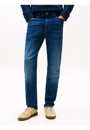 Homme portant un jean bleu foncé, des baskets beige et un pull texturé bleu foncé avec les mains dans les poches, debout sur un fond clair.