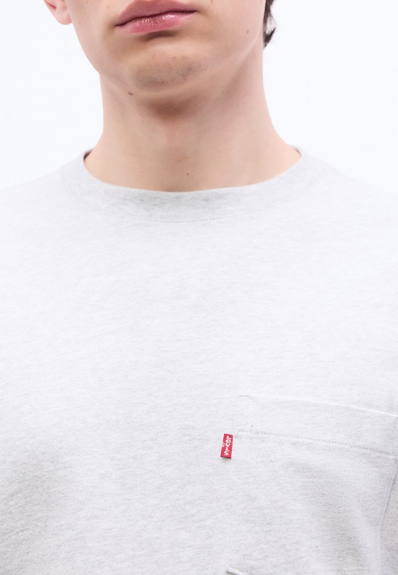 Primo piano di una persona che indossa una camicia grigio chiaro con una tasca sul petto e una piccola etichetta rossa con il logo Levi's sulla tasca.