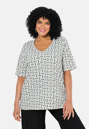Musteriertes V-Ausschnitt-Oberteil in Weiß mit schwarzem Zickzackdesign. Weicher Stoff, kurze Ärmel, entspannter Schnitt, kombiniert mit schwarzen Hosen.