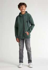 Zelená mikina na zip s přední kapsou a nášivkou s logem, nošená se šedými džíny. Oblečení doplňují bílé tenisky.