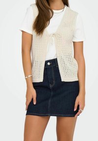 Beige knitted vest with tie-front closure over a white shirt, paired with a dark denim skirt. The vest features open weave and no sleeves.