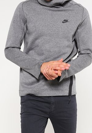 Personne portant un sweat gris Nike avec un zippé sur le côté, les mains jointes devant, associée à un pantalon sombre.
