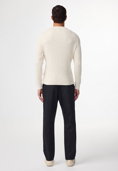 Beiger gerippter Pullover mit langen Ärmeln, figurbetontem Design und Rundhalsausschnitt, kombiniert mit schwarzen Hosen und hellen Schuhen, von hinten betrachtet.