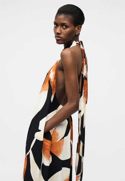 Γυναίκα με σκούρο δέρμα και κοντά μαλλιά, φορώντας αμάνικο, ανοιχτό φόρεμα στην πλάτη με μεγάλα πορτοκαλί και λευκά ανθισμένα σχέδια σε απλό φόντο.