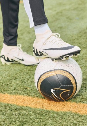 Biała korka na czarno-białej piłce nożnej spoczywającej na sztucznej trawie obok pomarańczowej linii, widoczne czarne spodnie i białe skarpetki.