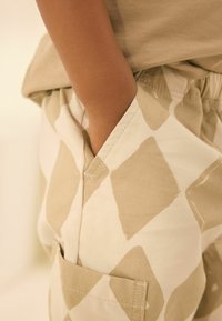 Beige gemusterte Baumwollshorts mit einem Diamantdesign und einem elastischen Bund, ausgestattet mit seitlichen Taschen und sichtbaren Nähdetails.