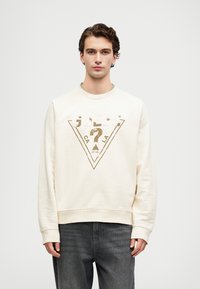 Ung mann iført en kremfarget hettegenser med en blek Guess-logo og falmede svarte jeans, stående mot en enkel lys bakgrunn.