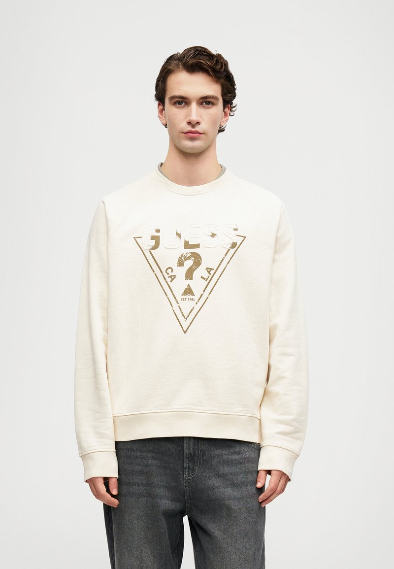 Ung mann iført en kremfarget hettegenser med en blek Guess-logo og falmede svarte jeans, stående mot en enkel lys bakgrunn.