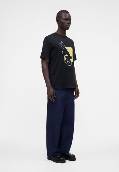 Homme portant un t-shirt noir avec un graphisme de lapin sur fond jaune, un pantalon ample bleu foncé et des chaussures noires, debout sur un fond blanc uni.