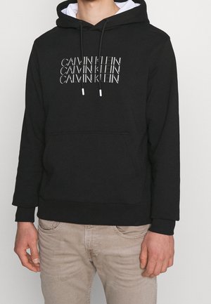 Sudadera negra con el texto "CALVIN KLEIN" en blanco en un patrón repetido. Cuenta con un bolsillo frontal y una capucha con cordón. Material de mezcla de algodón.