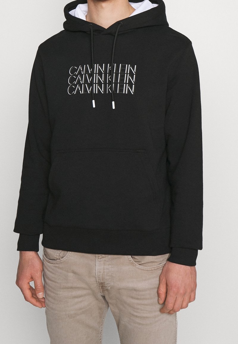 Felpa nera con testo bianco "CALVIN KLEIN" in un motivo ripetuto. Presenta una tasca a marsupio frontale e cappuccio con cordini. Materiale in misto cotone.