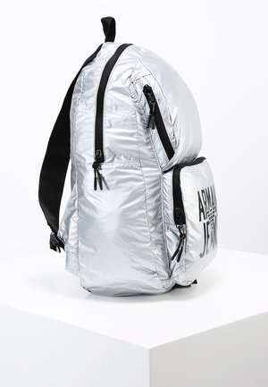 Sølv skinnende rygsæk med sorte lynlåse, en forlomme mærket "Armani Jeans" og sorte justerbare skulderstropper på en hvid piedestal.