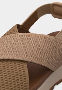 Gros plan sur une bride de sandale en tissu beige tissé et une bride rembourrée à la cheville sur une semelle marron et une semelle blanche.