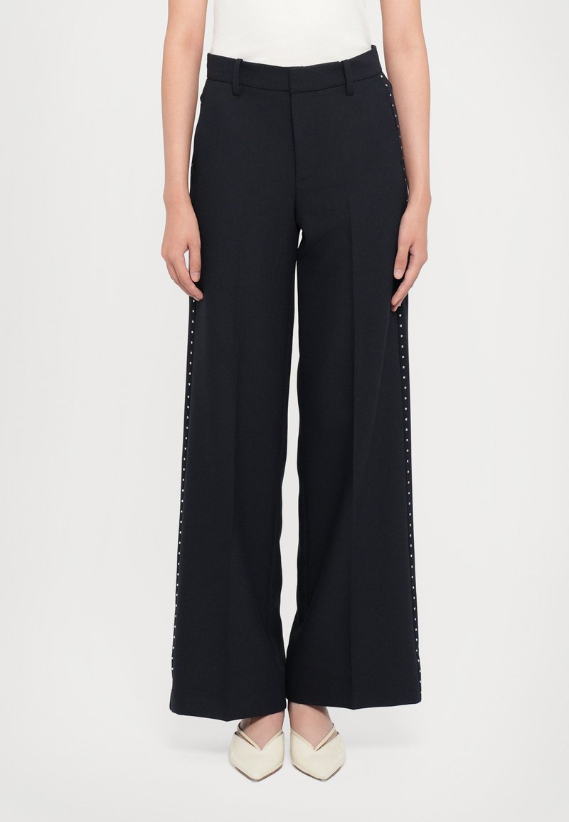 Wide-leg black trousers with side studs, tailored waist, and a smooth fabric finish. Paired with light-colored pointed-toe shoes.
