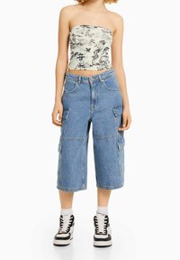 Žena má na sobě bílé crop top bez ramínek s potiskem černého draka a kaligrafie, široké denimové cargo kalhoty do poloviny stehen a černobílé tenisky.