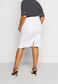 Woman wearing a black and white striped shirt, white knee-length skirt with back slit, and black high-heeled sandals, standing against white wall.