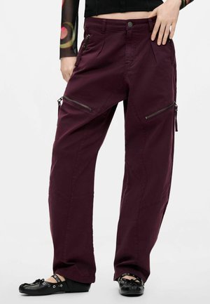 Person trägt weite bordeauxrote Cargohosen mit Reißverschlusstaschen, ein schwarzes gemustertes Langarmshirt und schwarze Ballerinas.