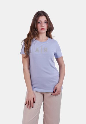 Donna con capelli castani lunghi, che indossa una maglietta lilla chiaro A|X Armani Exchange e pantaloni beige, in piedi davanti a uno sfondo chiaro e semplice.