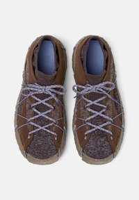 Hnědé venkovní tenisky s texturovanou síťovinou a vzorovanými tkaničkami, zachyceny zhora na bílém pozadí.