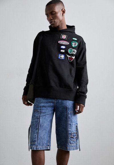 Black hooded sweatshirt with multiple colorful patches, paired with blue denim shorts featuring side pockets and a zipper detail.