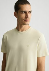Kurzärmliges T-Shirt mit Rundhalsausschnitt aus leichtem, beigem Baumwollstoff; verfügt über ein dezent geprägtes Logo auf der Brust; glatte, leichte Textur.