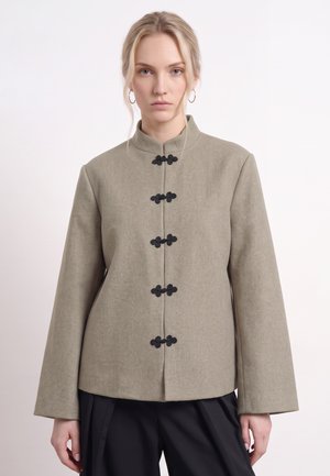 Une femme porte une veste taupe à col montant avec des brandebourgs noirs, des créoles et un pantalon noir sur un fond uni.