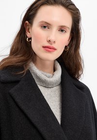 Manteau en laine noir avec de larges revers sur un col roulé gris clair. Présente un tissu texturé et des boucles d'oreilles en or rondes.