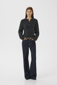 Femme se tenant debout avec les mains dans les poches, portant un pull boutonné gris foncé, une chemise à col large, un jean évasé noir, et une ceinture marron.