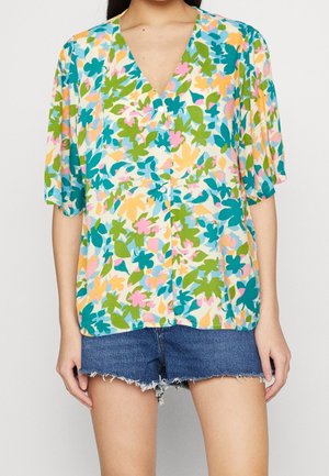 Femme portant un chemisier à manches courtes avec des boutons, avec un motif floral coloré vert, bleu, rose et jaune, et un short en jean effiloché.