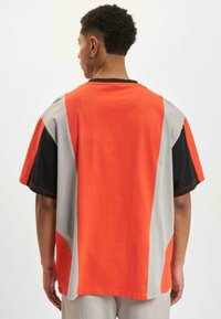 Tricou din bumbac cu blocuri de culoare, având secțiuni portocalii, gri și negre, cu mâneci scurte și o croială lejeră. Detalii de cusătură contrastante vizibile.