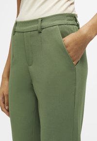Pantalon vert à coupe ajustée, avec une texture lisse, des poches latérales et une ceinture froncée.