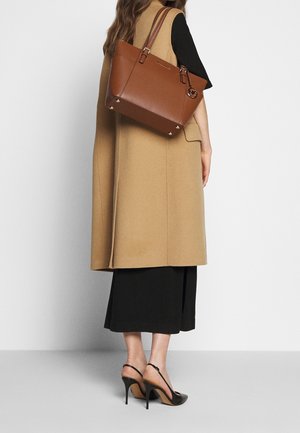 Femme portant un manteau sans manches couleur chameau, une robe noire, des escarpins noirs à talons ouverts, portant un grand sac fourre-tout en cuir marron sur son épaule.