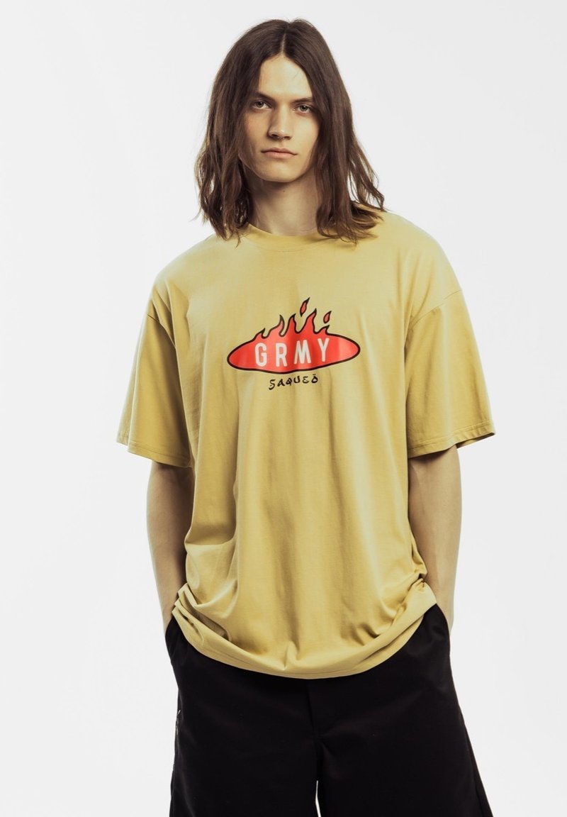 Camiseta de algodón beige con mangas cortas, que presenta un diseño de llamas rojas y el texto "GRMY SAUCE" en letras negras en negrita.