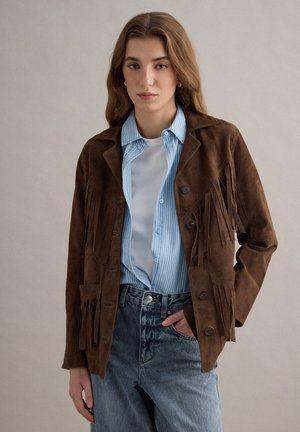 Jonge vrouw die een bruine suède jas met franjes draagt over een blauw gestreept overhemd en een wit topje, met high-waisted verweerde spijkerbroeken, staand met haar hand in haar zak.