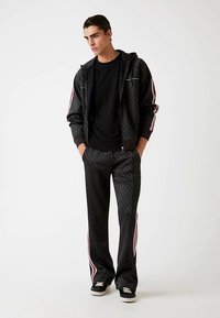 Sort træningsdragt i tekstureret stof med en hættetrøje med hvide striber og matchende bukser med sidelommer. Sorte sneakers.