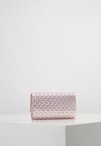 Pochette rectangulaire rose avec un motif en damier texturé composé de petits clous en relief, posée sur une surface blanche devant un fond uni.