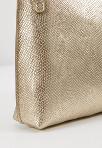 Zlatá metalická kabelka s texturou hadí kůže. Hladký povrch se strukturovaným tvarem a zesílenými okraji.