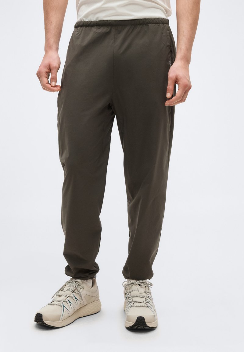 Homme portant un pantalon taille élastique olive foncé et des chaussures de sport blanches, debout les mains détendues le long du corps devant un fond uni.