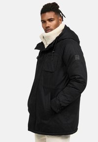 Parka negra con capucha, que presenta un collar de forro polar blanco texturizado, bolsillos con cremallera y un logo bordado en la manga.