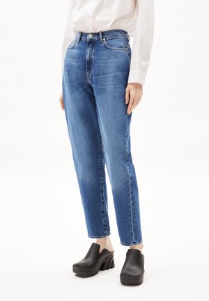 Personne portant un jean droit bleu, une chemise blanche à manches longues et des chaussures plateformes noires sans lacets, debout devant un fond blanc.