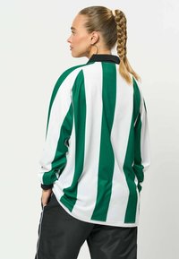 Femme avec une queue de cheval tressée portant une chemise à manches longues rayée verticalement verte et blanche et un pantalon noir, debout, les mains dans les poches.