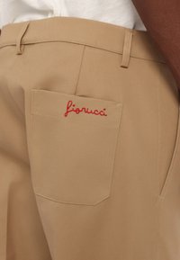 Beezhased püksid, millel on tagaosal tasku ja punane brodeeritud "fioruci" logo, valmistatud siledast ja vastupidavast kangast, millel on nähtavad õmblusdetailid.
