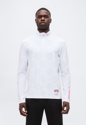 Jeune homme debout, portant un haut blanc à manches longues avec une fermeture éclair au quart et des motifs géométriques subtils, ainsi qu'un pantalon noir sur un fond uni.